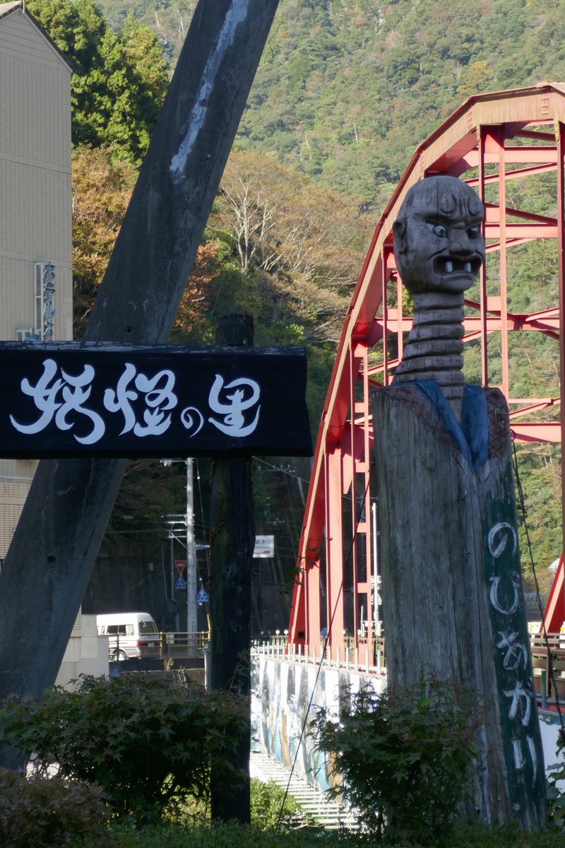 川沿いの道に立つ、首の長い木彫りの妖怪像