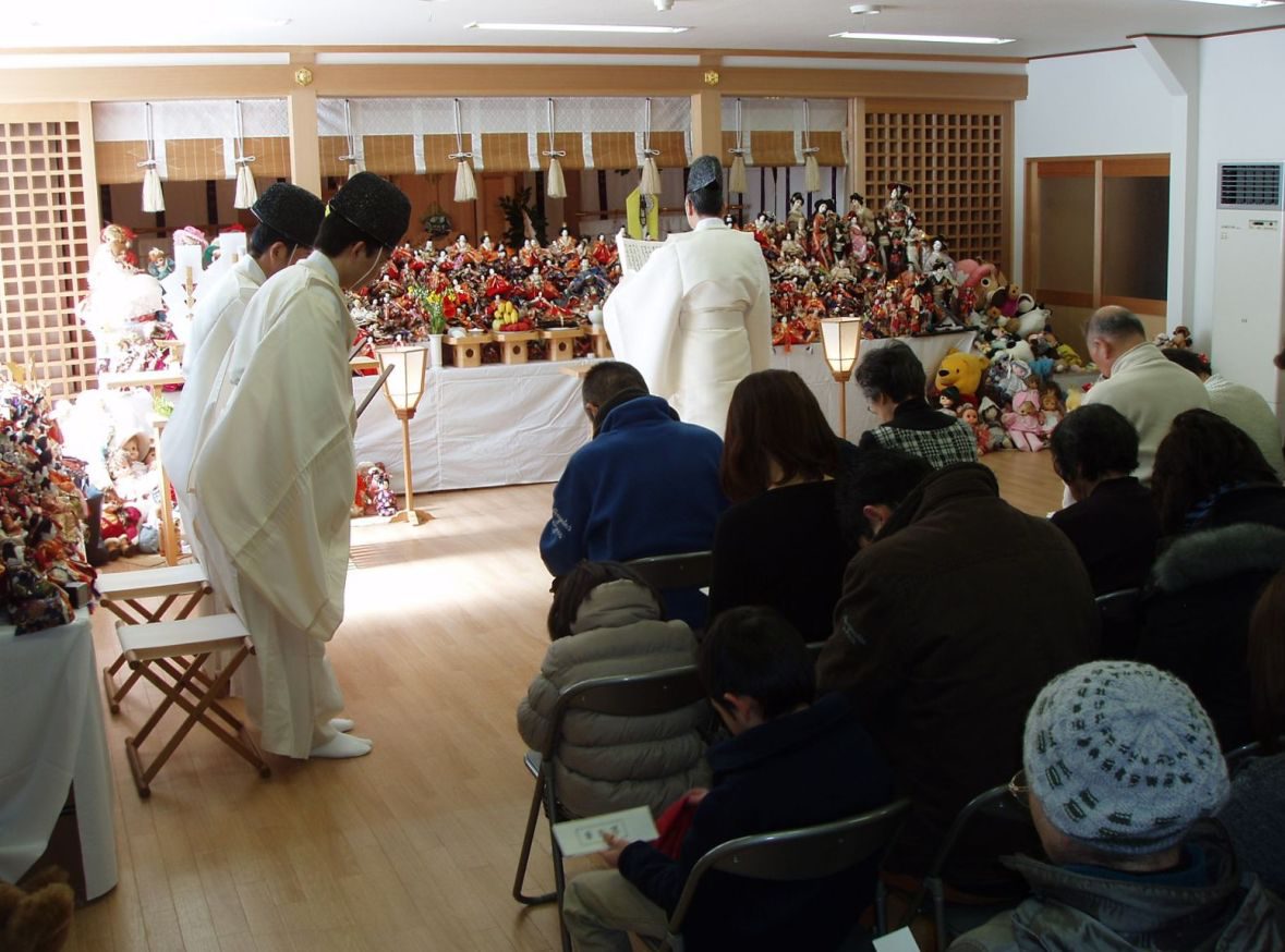 祭壇にたくさんの雛人形やぬいぐるみが並び、白い装束の神職の前に人々が座っている
