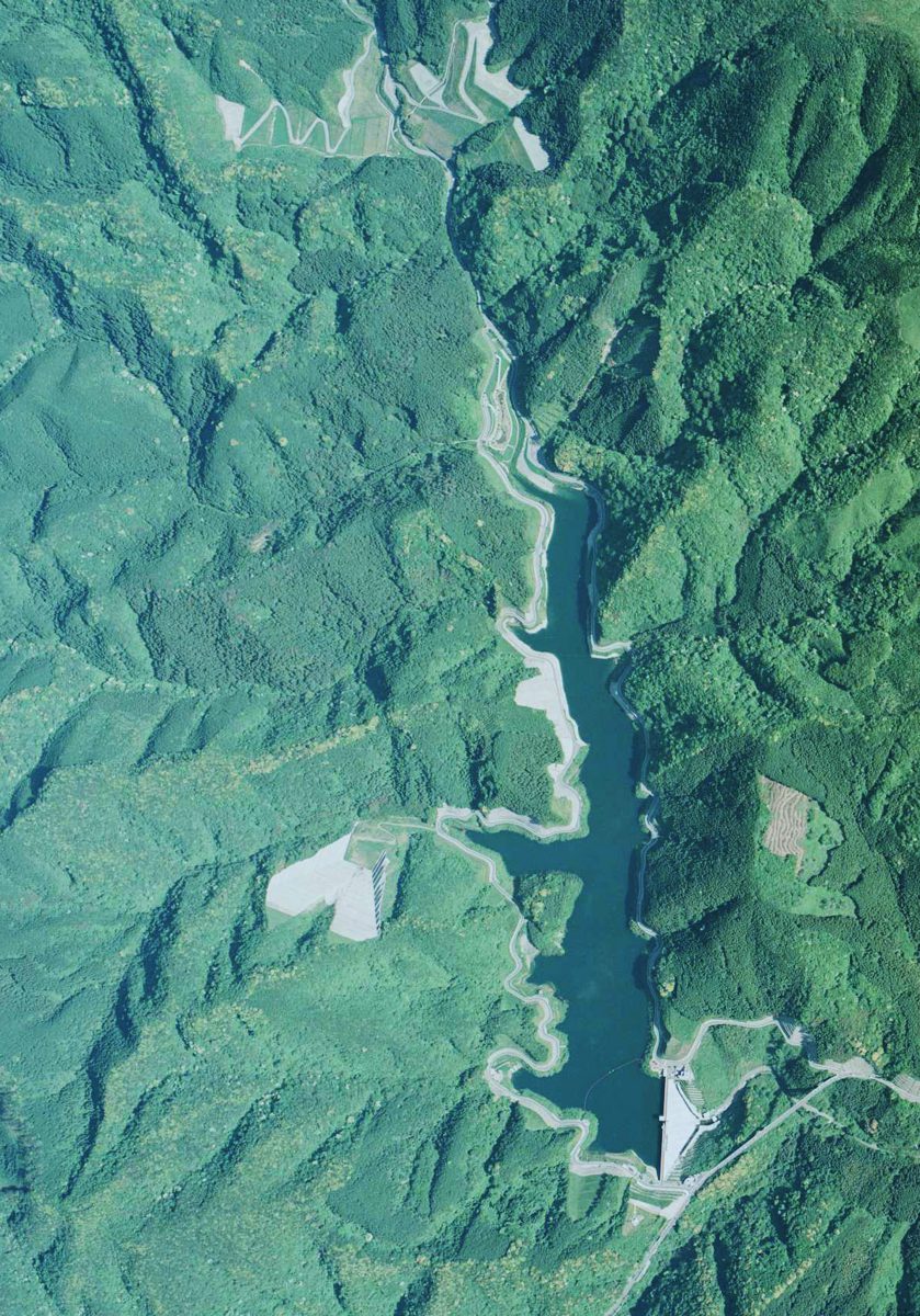 カラーの空中写真。山に囲まれた谷に、細長いダム湖が水をたたえている