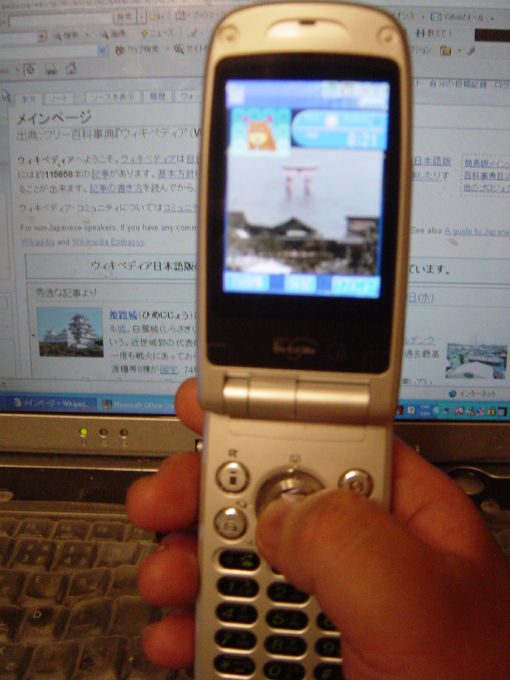 手に持った折りたたみ式の携帯電話。画面に神社の鳥居の映像が映っている