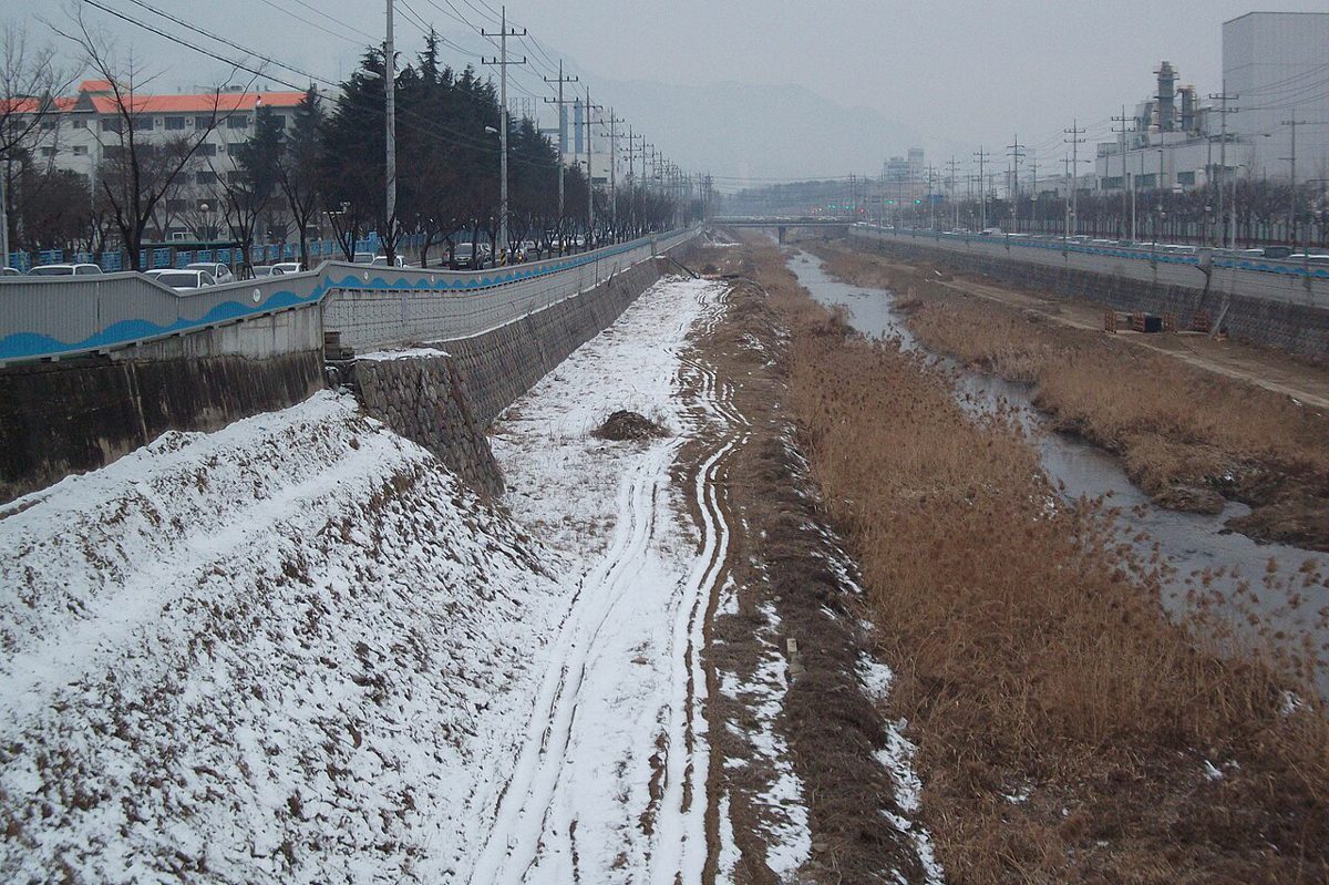 雪の残る土手と枯れ草の間を流れる川。両岸に道路と工場の建物が並ぶ