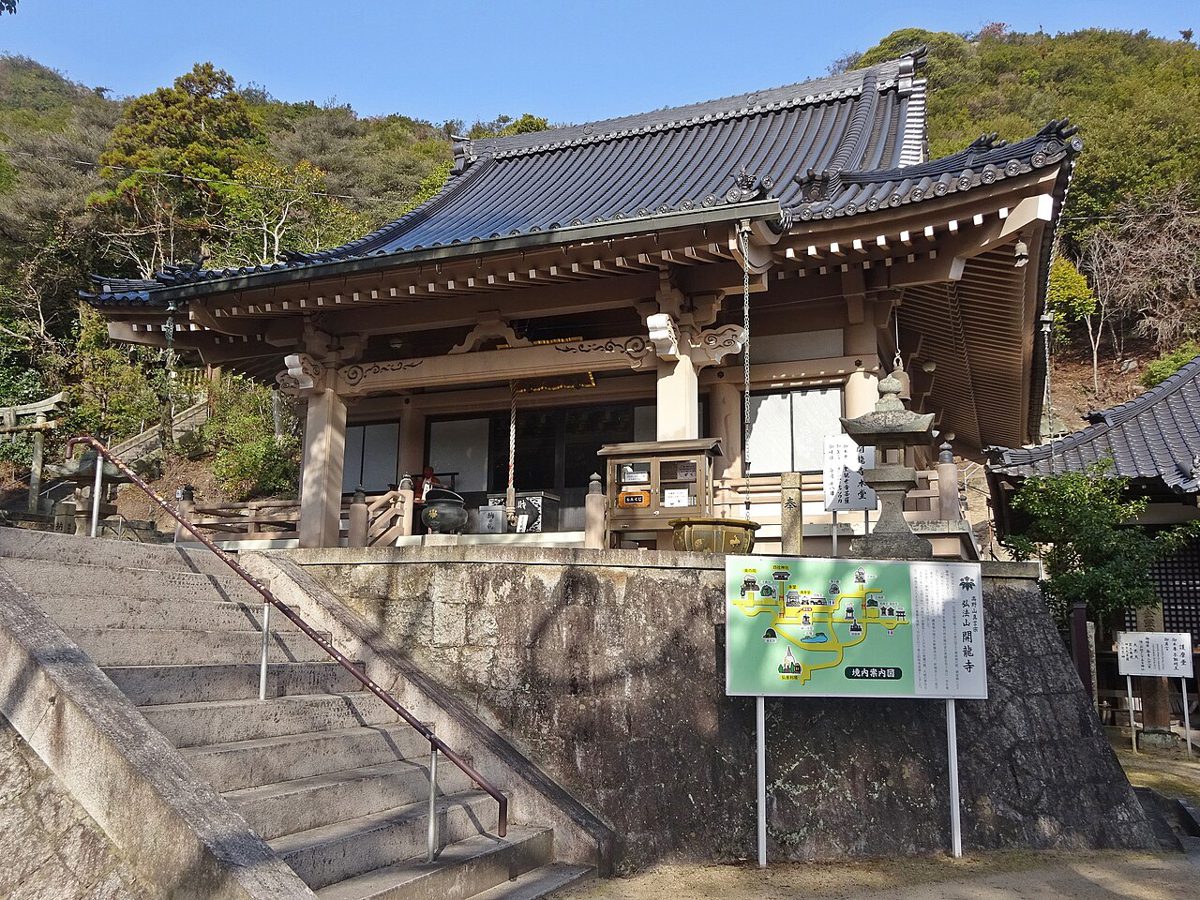 石段の上に建つ、反り屋根の寺の本堂。手前に境内の案内図の看板が立つ
