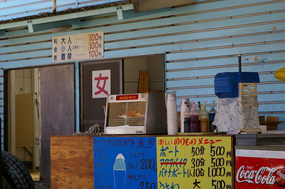 トタン張りの海の家の店先。かき氷やボート貸し出しの料金表が並ぶ