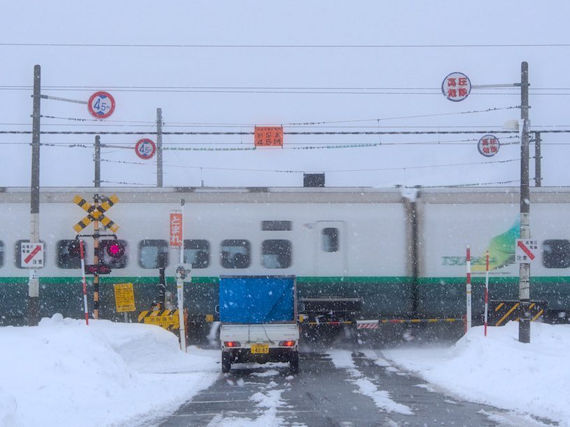 雪が降る中、踏切の前で軽トラックが止まり、その向こうを新幹線が通過している